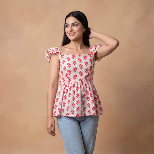 A woman modeling a white off-the-shoulder peplum top with a red floral print, paired with light blue jeans, against a warm beige studio backdrop.