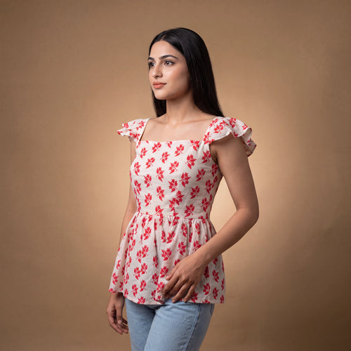 A woman modeling a sleeveless white peplum top with a red floral pattern, ruffled cap sleeves, and a square neckline, paired with light blue jeans against a warm brown studio backdrop.